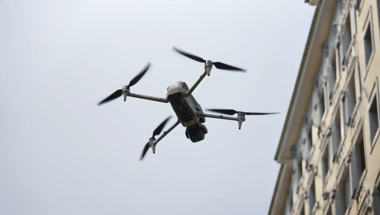 Inspection drone hovering beside the facade of an older multi-storey building, seen from below against an overcast sky